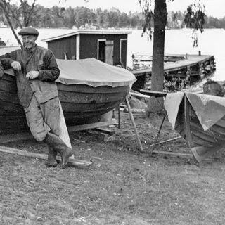Äldre svartvit bild av en man som lutar sig mot en träbåt på land vid varvet