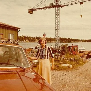 Äldre färgbild av en kvinna vid en bil på varvsplanen, med en lyftkran i bakgrunden